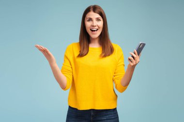 Heyecanlı, mutlu, gülümseyen orta yaşlı kadın cep telefonu tutuyor, mobil uygulama kullanıyor, kameraya bakıyor, mavi arka planda tek başına duruyor. Çevrimiçi alışveriş kavramı, sosyal medya