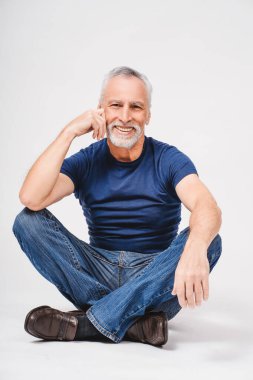 Beyaz arka planda izole edilmiş kameraya bakarak stüdyoda yerde oturan, tişörtlü, gülümseyen gri saçlı adamın portresi. Dikey. Reklam kavramı
