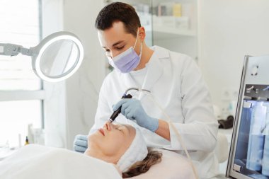 Profesyonel erkek, kozmetik uzmanı, yaşlı kadın üzerinde mezoterapi uyguluyor, hasta su yüzüne mikrodermabrüksiyon yaptırıyor. Yaşlı kadın estetik ameliyat oluyor. Kozmetoloji kavramı