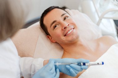 Gülümseyen, genç bir adam, modern kozmetik kliniğinde yatan bir hasta, kozmetik uzmanı ile konuşuyor, kozmetik prosedürleri, güzellik enjeksiyonu, gençleştirme. Kozmetoloji kavramı