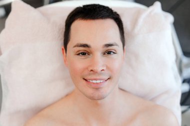 Yakışıklı, gülümseyen bir adam, kozmetik kliniğinde manken, yalan söylüyor, kameraya bakıyor, kapatıyor. Cilt bakımı kavramı, kozmetoloji