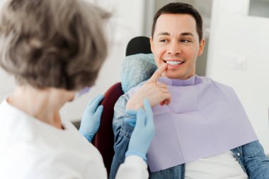 Genç ve çekici bir erkek hasta dişlerini göstererek dişlerini gösteriyor. Ağrısı var. Modern diş kliniğindeki profesyonel dişçi doktoruyla konuşuyor. Diş bakımı kavramı, sağlık hizmetleri