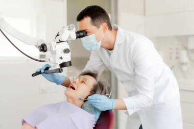 Genç dişçi doktor, diş mikroskobuyla diş tedavi eden adam, yaşlı çekici kadını kontrol eden, emekli, modern dişçilikte çalışan adam. Profesyonel ekipman kavramı, diş bakımı