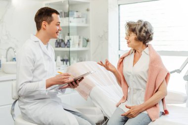 Profesyonel güzellik doktoru, beyaz önlüklü adam, modern klinikte oturan yaşlı kadın, hasta ile konuşuyor. Sağlık ve iletişim kavramı