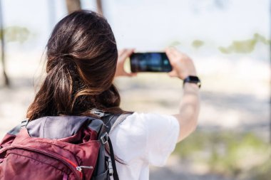 Sırt çantalı gezgin kadın kayalık sahilde duruyor ve akıllı telefondan yeşil tepelerin ve dağların fotoğrafını çekiyor. Yürüyüş konsepti
