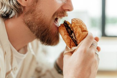 Sakallı bir adam elinde bir burger tutuyor ve büyük bir ısırık alıyor. Peynirli ve taze sebzeli sulu köfte çöreklerin arasında görünür.