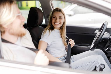 Genç bir kadın emniyet kemerini bağlarken gülümsüyor, bir arabanın yolcu koltuğunda genç bir adamla oturuyor.