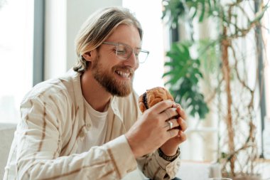 Sakallı ve gözlüklü genç adam evde kruvasanın keyfini çıkarıyor, mutlu bir şekilde gülümsüyor. Yeşil bitkiler ve doğal ışıkla çevrili tatmin edici bir ısırık alır.