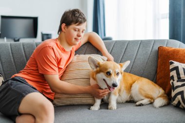 Down sendromlu gülümseyen genç çocuk evdeki kanepede oturmuş evcil Galler 'li Corgi Pembroke' una sarılıyor. Arkadaşlık, aşk, hayvan konsepti.