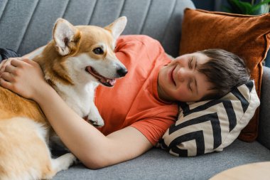 Down sendromlu mutlu bir genç evdeki kanepede dinleniyor, evcil köpeğine sarılıyor. Hayvan konsepti, rahatlatıcı. Hayvan sevgisi konsepti