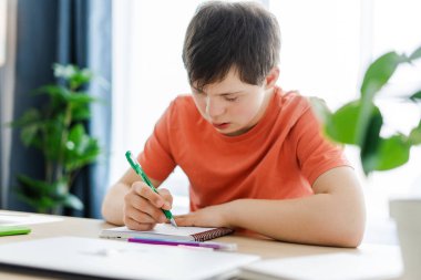 Down sendromlu genç, ödevlerini evde yapıyor, masasına not defteri yazıyor, not alıyor. Eğitim kavramı