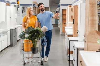 Gülümseyen evli genç bir çiftin portresi. Tesisatçı dükkanındaki banyo mobilyalarına bakıyorlar. Alışveriş arabası kullanıyorlar.