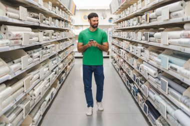 Cep telefonu tutarken hırdavatçıdaki raflardan duvar kağıtlarını seçen mutlu sakallı bir gencin portresi. Alışveriş konsepti