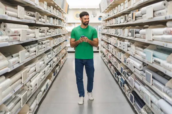 Cep telefonu tutarken hırdavatçıdaki raflardan duvar kağıtlarını seçen mutlu sakallı bir gencin portresi. Alışveriş konsepti