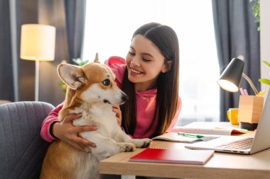 Genç kız, genç kız rahat bir oturma odasında Corgi köpeğine sarılırken gülümsüyor, evde ders çalışmaya ara veriyor. Hayvan konseptine bayılırım.