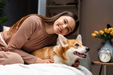 Gülümseyen ergen, tanıdık eşyalarla çevrili rahat bir yatak odasında Corgi 'sine sarılıyor. Neşeli dostluklarını ve memnuniyetlerini yakalıyor. Hayvan konseptine bayılırım.