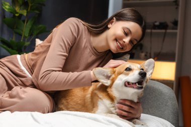 Genç bir kız, evdeki bir yatakta dinleniyor, sevimli Corgi köpeğiyle eğleniyor, samimi bir arkadaşlık ve aşk sahnesi yaratıyor.
