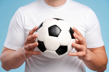 Beyaz tişörtlü kararlı bir sporcu elinde futbol topu tutuyor ve oyuna olan tutkusunu gösteriyor. Yoğun odaklanma, klasik renkler ve mavi arka plan spor heyecanını vurguluyor