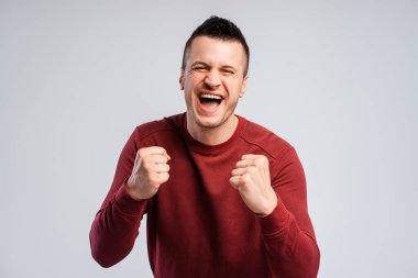 Yakından gülümseyen genç hayran kameraya bakıyor ve yumruklarını sıkıyor ve evet diyor. Kazanma ve spor konsepti. Gri arkaplanda izole edilmiş duruyor.