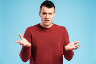 Tatminsiz, üzgün, mavi arka plandaki kırmızı kazaklı adam hayal kırıklığı içinde ellerini kaldırır.