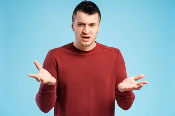 stock image Dissatisfied, upset man in red sweater on blue background throws up his hands in frustration