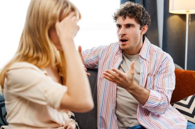 Genç adam, sıcak bir oturma odasında bir kadınla ateşli bir tartışma sırasında öfkeyle el kol hareketi yapıyor. Duygusal gerginliği yansıtıyor ve ilişkilerde etkili iletişimin önemini vurguluyor.