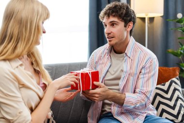 Heyecanlı bir adam evdeki oturma odasında şaşkın bir kadına hediye veriyor. Çekici erkek kırmızı hediye kutusu tutuyor. Kutlama konsepti