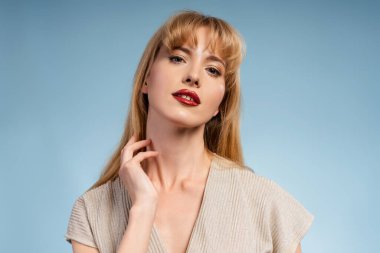 Kırmızı rujlu ve göz alıcı bluzu olan kendine güvenen genç bir kadın mavi arka planda poz veriyor, eliyle boynuna dokunuyor.