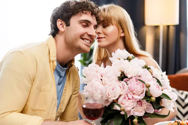 stock image Man surprises woman with peonies and wine in a cozy living room, sharing a joyful, loving moment. The gesture adds tenderness to their celebration, symbolizing affection and elegance