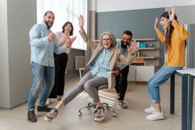 Çeşitli ofis çalışanları gülüyor ve olgun bayan patronlarını ofisin karşısındaki sandalyeye oturturken eğleniyorlar. Takım oluşturma kavramı