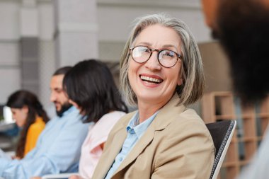 Olgun, neşeli bir iş kadını iş arkadaşlarıyla canlı bir toplantı yapıyor, uyumlu bir iş yerinde dizüstü bilgisayarlar ve bilgisayarlarla güven ve takım çalışması sergiliyor. Takım çalışması konsepti