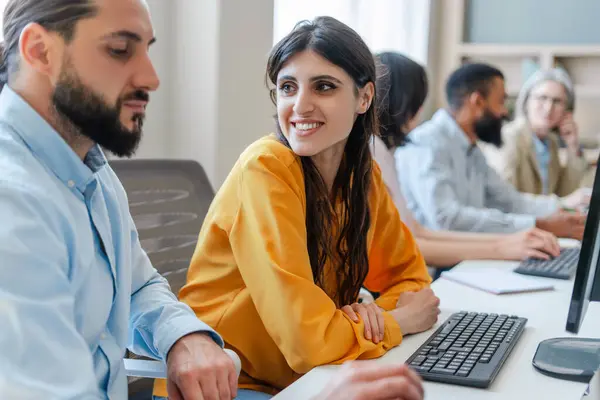 stock image Happy young businesswoman works with a colleague in a busy office, showcasing teamwork and collaboration among diverse businesspeople using technology