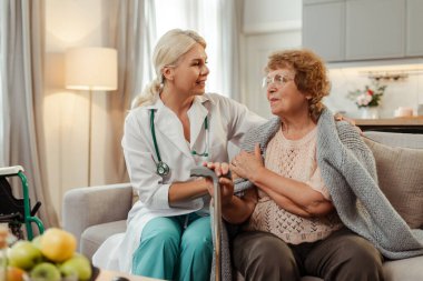 Değer veren kadın doktor evde yaşlı bir kadına destek oluyor, empati ve güven iletiyor. Olay yeri, ev sağlık hizmetlerinde iletişimi ve kaliteli tıbbi bakımı teşvik etmektedir.