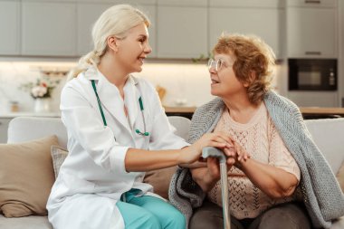 Yaşlı bir kadının yanındaki kanepede oturan gülümseyen kadın doktor baston tutuyor, ikisi de gülümsüyor ve sohbet ediyorlar. Sağlık, destek, tedavi kavramı