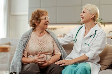 Beyaz önlüklü ve steteskoplu kadın doktor evde gülümseyen yaşlı bir hastayı dinliyor ve onunla konuşuyor. Sağlık hizmetlerinde rahatlığın önemini gösteriyor. İletişim konsepti, destek
