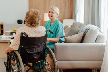 Neşeli hemşire bir koltukta oturuyor, tekerlekli sandalyedeki yaşlı bir kadınla konuşuyor, aydınlık ve rahat bir ev ortamında. Toparlanma kavramı
