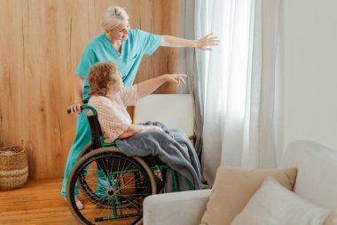 Pencerenin yanında yaşlı bir kadınla ilgilenen genç hemşire dışarıda gülümseyen kadını işaret ediyor. Toparlanma kavramı