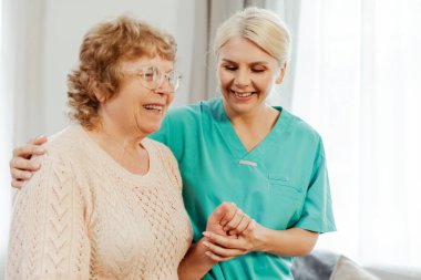 Gülümseyen, mutlu doktor, yaşlı kadının elini tutarak evde vakit geçiriyor. Rehabilitasyon, iyileşme kavramı