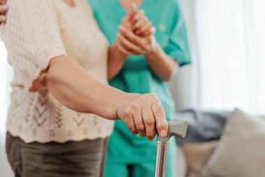 Hemşire arka planda dikilirken baston kullanan yaşlı bir kadın. İyileşme, destek kavramı