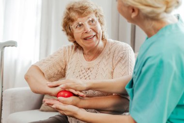 Hemşire, evde iyileşme sürecinde stres topu kullanan mutlu bir yaşlı kadına yardım ediyor. Kadın bir kanepede oturuyor ve topu elinde tutuyor. Rehabilitasyon kavramı