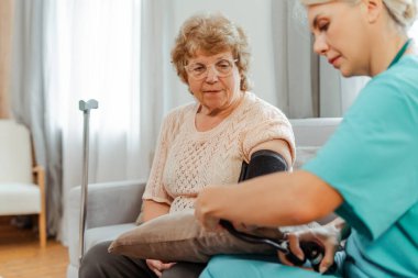 Bakıcı kadın doktor, ev ziyareti sırasında yaşlı bir hastanın kan basıncını gözlemler ve sağlık hizmetlerine duyulan güven ve empatiyi yansıtır.