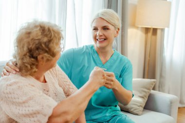 Gülümseyen hemşire, yaşlı bir kadını evinde ziyaret ediyor. Yumruk tokuşturarak ilgi ve destek veriyor. Şefkat ve güven gösteriyor.