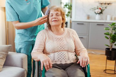 Hemşire, yaşlı ve mutlu bir kadının saçını nazikçe tarar. Tekerlekli sandalyede oturur ya da yardım alarak yaşar.