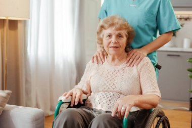 Hemşire evde tekerlekli sandalyede yaşlı bir kadına bakıyor, şefkat ve destek gösteriyor.