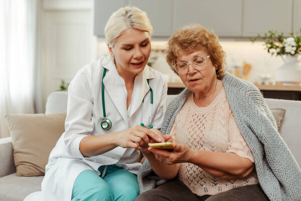 Улыбающийся доктор показывает пожилой женщине медицинское приложение во время домашнего визита, сидя на диване в уютной гостиной. Поддержка, концепция здравоохранения