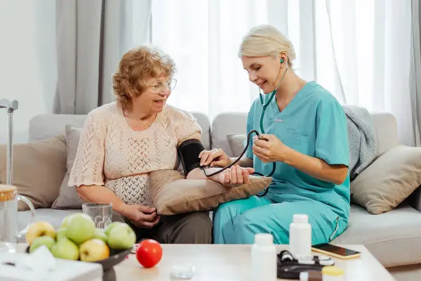 Önlük giymiş gülümseyen hemşire oturma odasındaki kanepede oturan neşeli, son sınıf öğrencisi bir kadının tansiyonunu ölçüyor. Sağlık hizmeti kavramı