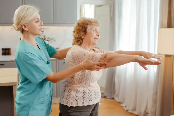 stock image Nurse helps a senior woman with arm stretches at home, promoting mobility and wellbeing. Smiling, they show the positive impact of healthcare on active aging