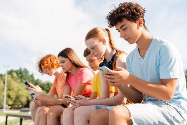 Cep telefonlu bir grup genç açık hava bankında sırada oturuyorlar. Teknoloji bağımlılığı kavramı