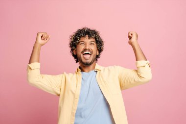 Heyecanlı genç adam pembe arka planda başarıyı kutluyor, yumruklarını kaldırıyor ve neşeyle bağırıyor, haz ve gurur saçıyor. Olumlu duygular ve coşku saçan kıvırcık saçlı.