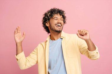 Kıvırcık saçlı genç adam pembe bir arka plan karşısında günlük kıyafetleriyle mutlu bir şekilde dans ediyor, neşe ve pozitiflik saçıyor. Sevinç ve neşe aktarımı için mükemmel.